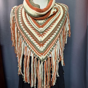 May include: A triangular crochet shawl featuring a striped pattern in cream, olive green, and burnt orange. The garment has a textured stitch design and is finished with long, dangling fringe tassels in matching colors along the bottom edge.
