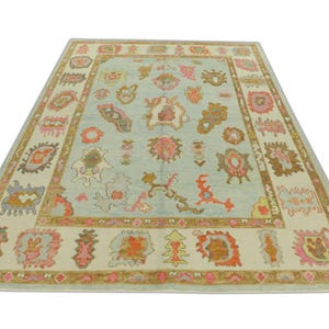 May include: A rectangular area rug with a light blue center and a cream-colored border. The rug features a repeating pattern of colorful, abstract floral and geometric shapes in shades of pink, orange, and brown.