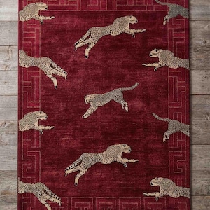 May include: A rectangular area rug in deep red featuring a repeating pattern of leaping cheetahs in tan and black. The rug is framed by a geometric Greek key border in a lighter red tone and is finished with cream-colored fringe on both ends.
