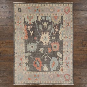May include: Rectangular area rug with a dark gray center and a light gray border. The rug features floral and abstract designs in shades of coral, blue, and cream. The rug is placed on a dark wood floor.