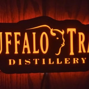 May include: Illuminated sign for Buffalo Trace Distillery. The sign is black with orange lettering and a buffalo silhouette. The words "BUFFALO TRACE" are at the top, and "DISTILLERY" is at the bottom.