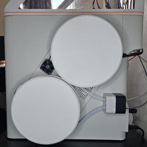 Puede incluir: Una impresora 3D blanca con dos grandes bobinas de filamento circulares montadas en el lateral. La máquina incluye un pequeño ventilador de refrigeración, un concentrador de filamento blanco y negro con dos tubos translúcidos y varios cables.
