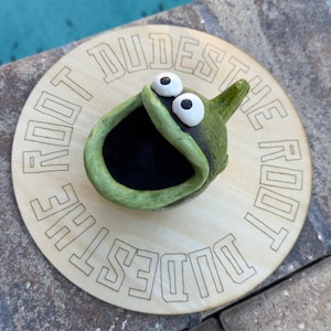May include: A whimsical, green ceramic sculpture with large, googly eyes and an open mouth, atop a wooden round sign. The sign is engraved with the words "DUDE THE ROOT" in a circular pattern. The sculpture has a black interior.