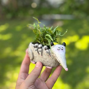 May include: A whimsical, hand-painted ceramic planter shaped like an opossum, with a long snout, big eyes, and small ears. The planter is filled with green foliage. The opossum has a light gray body, pink feet, and black ears. The planter is held in a hand.