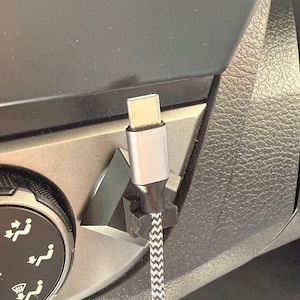 May include: A silver and black USB-C charging cable is plugged into a car's dashboard. The cable has a braided black and white pattern. The dashboard is black and grey.