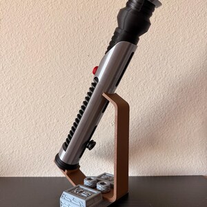 May include: A silver and black lightsaber prop with a red button, displayed on a brown stand. The stand has a detailed base and two small, gray cylinders. The lightsaber is angled, showcasing its intricate design.