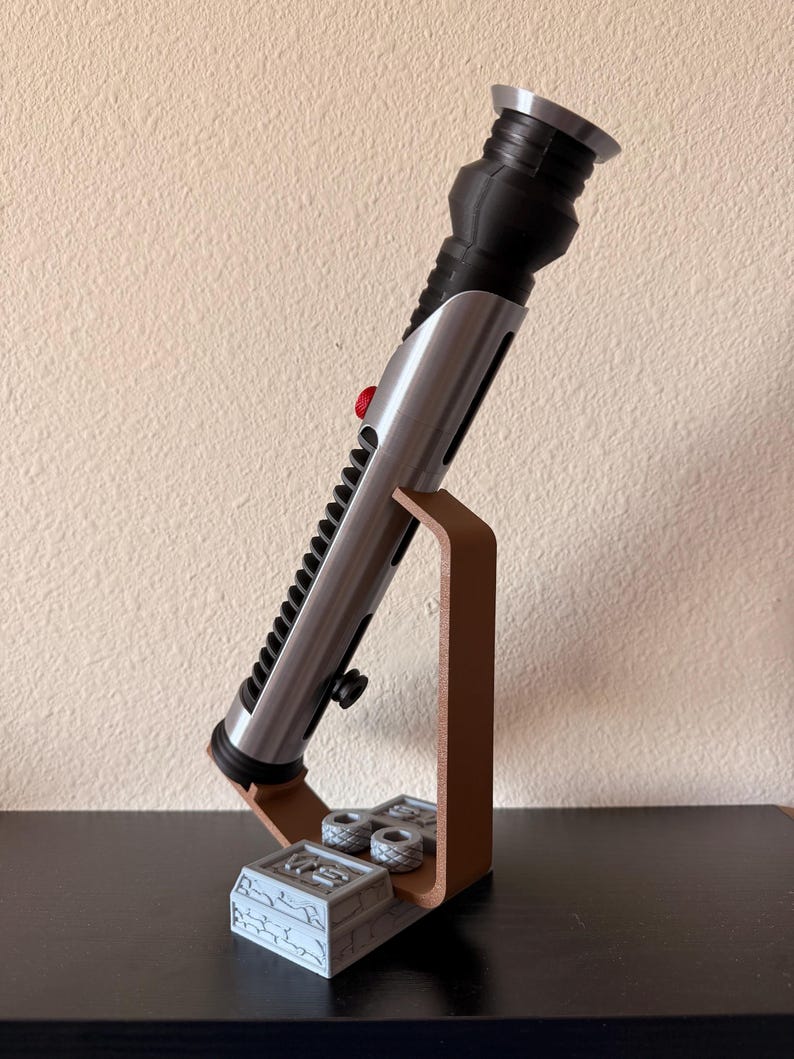 May include: A silver and black lightsaber prop with a red button, displayed on a brown stand. The stand has a detailed base and two small, gray cylinders. The lightsaber is angled, showcasing its intricate design.