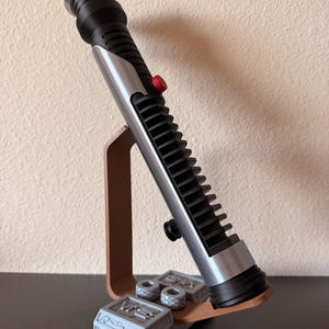 May include: A black and silver lightsaber prop with a red button, displayed on a brown stand. The stand also holds two small gray cylinders and a gray rectangular block with engraved details. The lightsaber is a replica of a popular science fiction weapon.