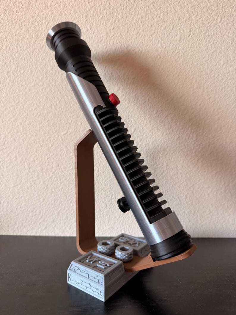 May include: A black and silver lightsaber prop with a red button, displayed on a brown stand. The stand also holds two small gray cylinders and a gray rectangular block with engraved details. The lightsaber is a replica of a popular science fiction weapon.