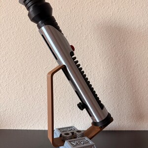 May include: A silver and black lightsaber prop, with a red button and black ridges, is displayed on a brown and gray stand. The stand has a rectangular base with engraved details and a curved support.
