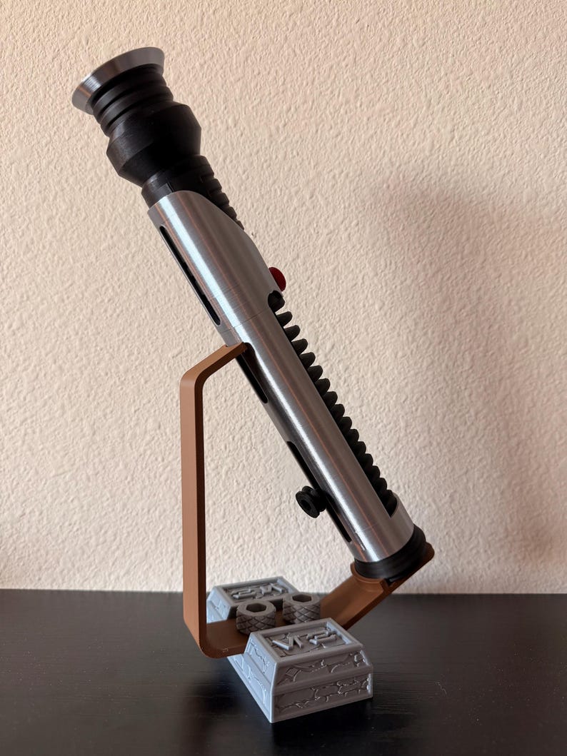 May include: A silver and black lightsaber prop, with a red button and black ridges, is displayed on a brown and gray stand. The stand has a rectangular base with engraved details and a curved support.