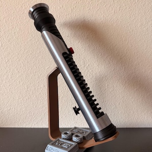 May include: A silver and black lightsaber prop with a red button, displayed on a brown stand. The stand also holds two small, gray, rectangular boxes with circular details. The lightsaber is angled, showcasing its intricate design.