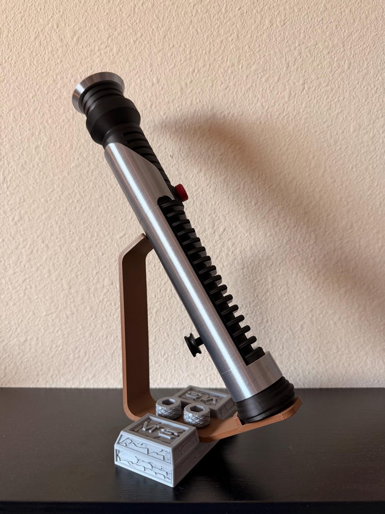 May include: A silver and black lightsaber prop with a red button, displayed on a brown stand. The stand also holds two small, gray, rectangular boxes with circular details. The lightsaber is angled, showcasing its intricate design.