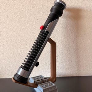 May include: A black and silver lightsaber prop with a red button, resting on a brown stand with a gray base. The lightsaber has a ribbed handle and silver accents. The stand is on a dark surface.