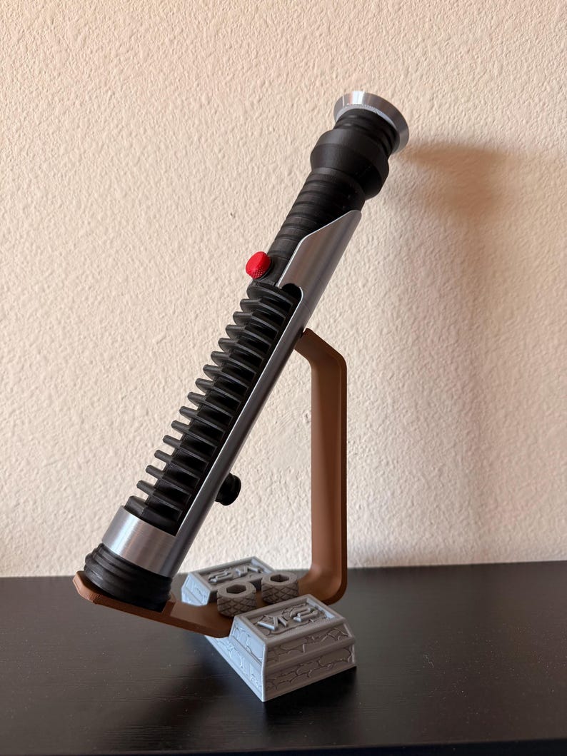 May include: A black and silver lightsaber prop with a red button, resting on a brown stand with a gray base. The lightsaber has a ribbed handle and silver accents. The stand is on a dark surface.