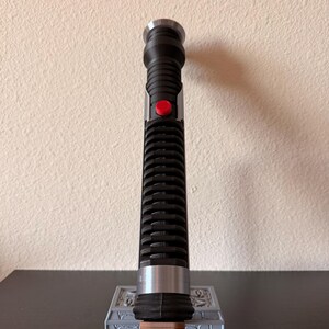 May include: A black and silver lightsaber with a red button, resting on a gray stand. The lightsaber has a ribbed handle and a silver emitter. The stand has a detailed design.