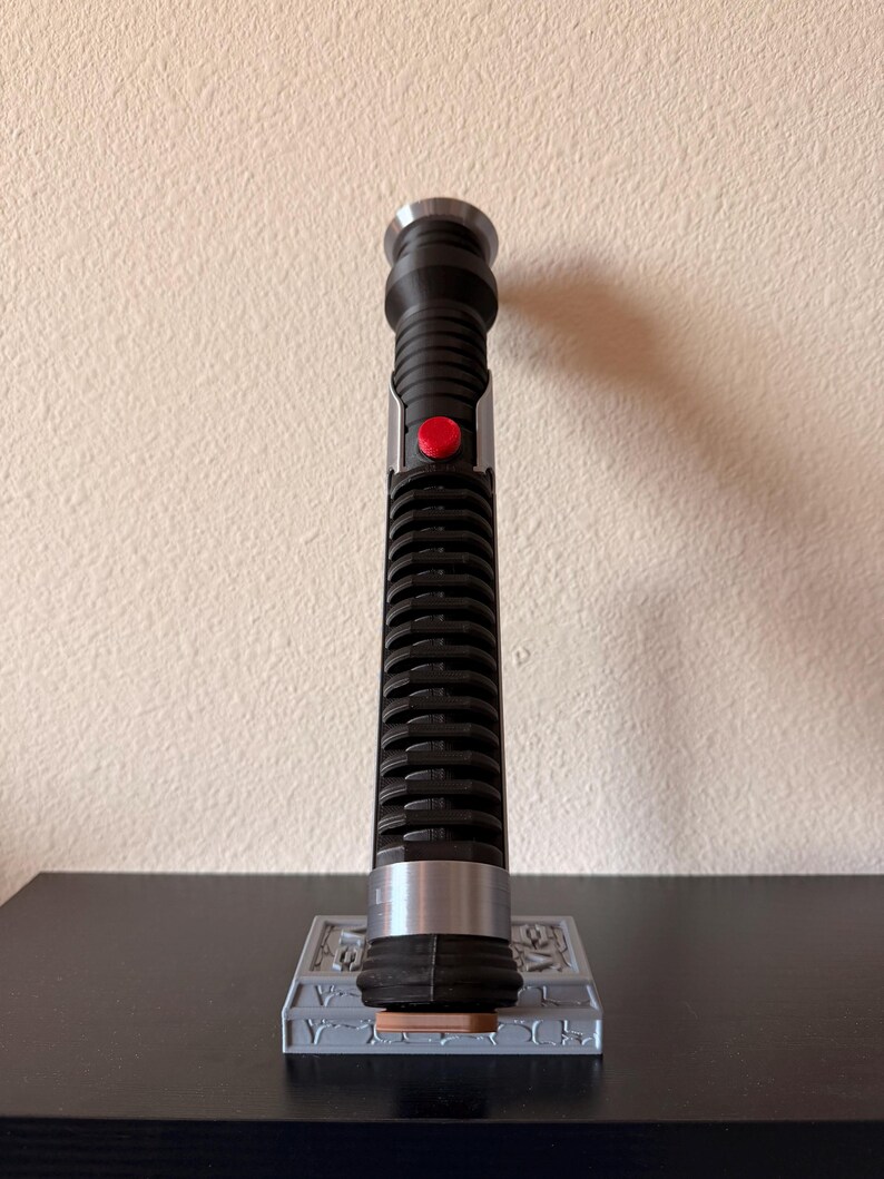 May include: A black and silver lightsaber with a red button, resting on a gray stand. The lightsaber has a ribbed handle and a silver emitter. The stand has a detailed design.