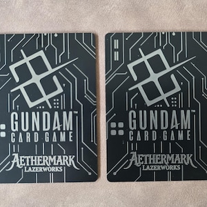 May include: Two black Gundam Card Game cards with silver circuit board designs. The cards feature the Gundam Card Game logo, Aethermark, and Lazerworks text. The background is a neutral color.