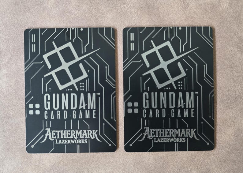 May include: Two black Gundam Card Game cards with silver circuit board designs. The cards feature the Gundam Card Game logo, Aethermark, and Lazerworks text. The background is a neutral color.