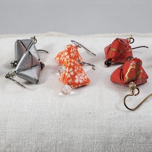 May include: A collection of handmade origami earrings in various colors and designs. The earrings feature folded paper in shades of orange, red, silver, and teal, with gold accents and beaded details. Some have a floral pattern.