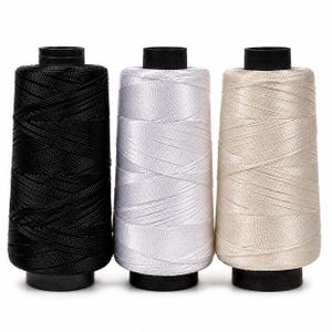 May include: Three cylindrical spools of thread arranged side by side. The left spool is black, the center spool is white, and the right spool is a light beige. Each spool features a black plastic core at the top and bottom with thread wound around.