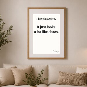 May include: A framed wall art print featuring the black text I have a system. It just looks a lot like chaos. with the brand name Ordiform in the bottom right corner. The print is displayed in a light wood frame against a neutral wall.