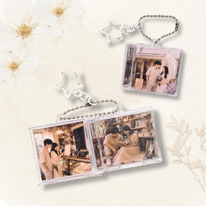 Puede incluir: Dos llaveros cuadrados de plástico transparente con diseño de caja de CD en miniatura. Cada llavero incluye una cadena de bolas plateada y un cierre en forma de estrella. Las inserciones muestran fotos de una pareja en interiores.