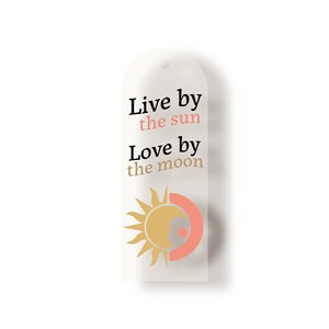 Può includere: Un'etichetta bianca con il testo "Live by the sun, Love by the moon". L'etichetta presenta un sole e una luna grafici in giallo, pesca e grigio. L'etichetta ha una parte superiore arrotondata e un piccolo foro per appendere.