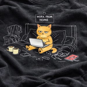 May include: Black t-shirt featuring a cartoon cat working on a laptop in bed. The cat has an annoyed expression. The words "WORK FROM HOME" are above the cat. Other items include coffee mugs, a phone, and a notebook.