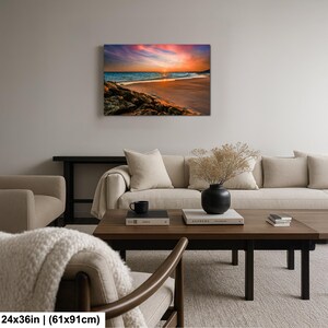 May include: A canvas print depicting a beach scene at sunset. The artwork showcases a vibrant sky with hues of orange, pink, and purple, reflecting on the ocean. The beach features golden sand and rocky areas. The dimensions are 24x36 inches.
