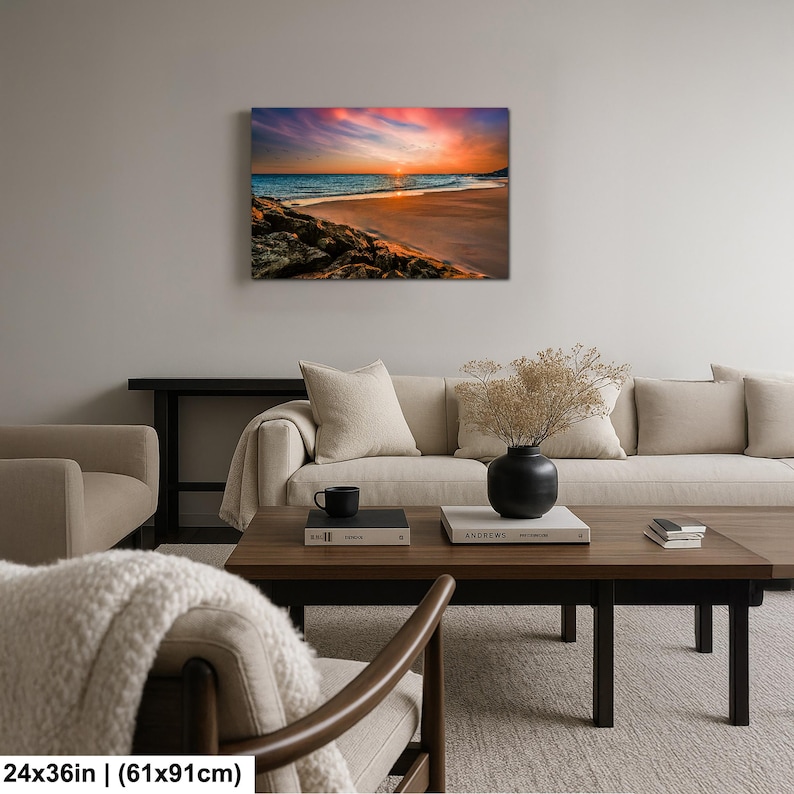 May include: A canvas print depicting a beach scene at sunset. The artwork showcases a vibrant sky with hues of orange, pink, and purple, reflecting on the ocean. The beach features golden sand and rocky areas. The dimensions are 24x36 inches.
