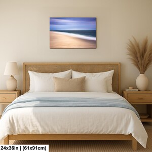 May include: A framed canvas print of a beach scene with a blurred effect, measuring 24x36 inches. The artwork depicts a sandy beach with the ocean and sky in shades of blue, beige, and white. The print is displayed above a bed with white bedding and a wooden headboard.