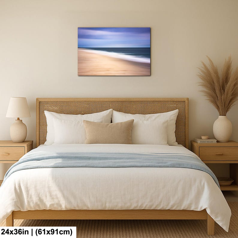 May include: A framed canvas print of a beach scene with a blurred effect, measuring 24x36 inches. The artwork depicts a sandy beach with the ocean and sky in shades of blue, beige, and white. The print is displayed above a bed with white bedding and a wooden headboard.