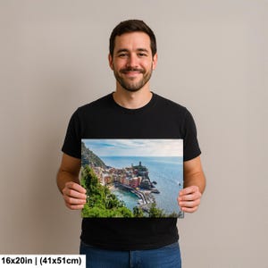 May include: A 16x20 inch (41x51cm) canvas print featuring a coastal village scene. The image shows colorful buildings nestled along a cliffside, with a lighthouse and boats in the sea. Lush green vegetation frames the village.
