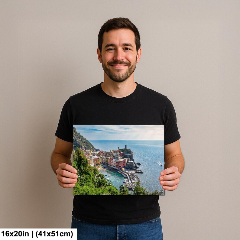 May include: A 16x20 inch (41x51cm) canvas print featuring a coastal village scene. The image shows colorful buildings nestled along a cliffside, with a lighthouse and boats in the sea. Lush green vegetation frames the village.