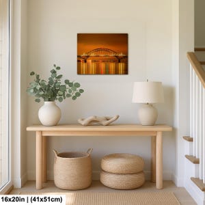 May include: A framed canvas print of a bridge at night, with lights reflecting in the water. The print is 16x20 inches (41x51cm). The print hangs above a wooden console table with a white vase, a lamp, and woven baskets.