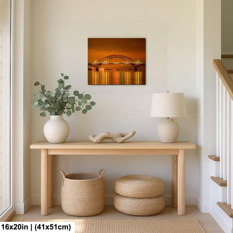May include: A framed canvas print of a bridge at night, with lights reflecting in the water. The print is 16x20 inches (41x51cm). The print hangs above a wooden console table with a white vase, a lamp, and woven baskets.
