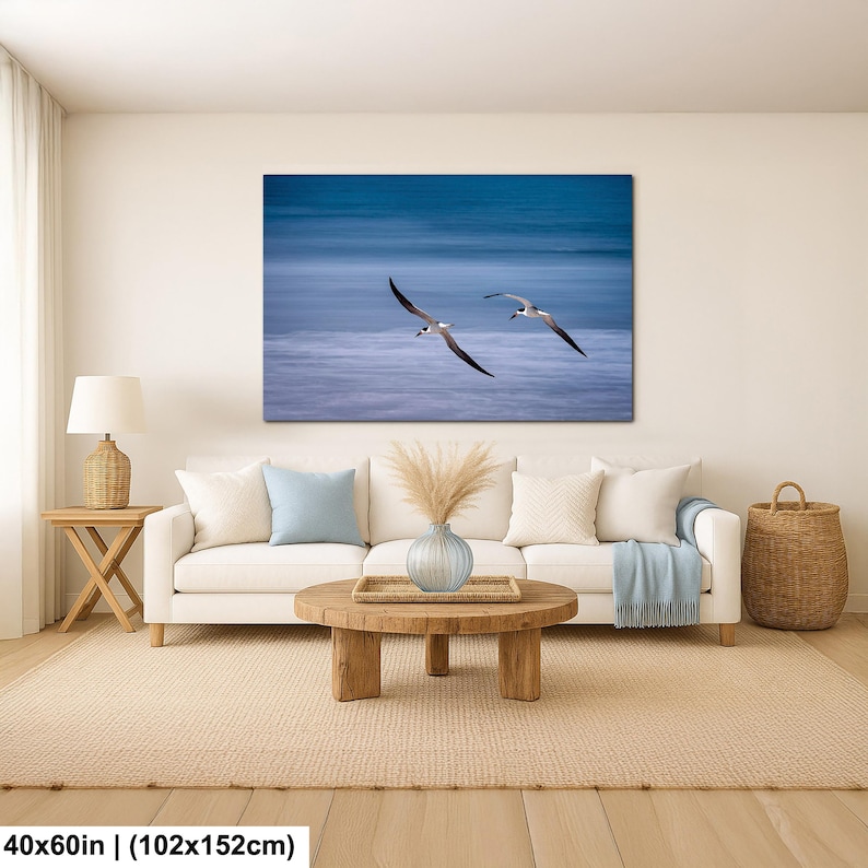 May include: A canvas print featuring two birds in flight over a blue ocean. The artwork is displayed above a white sofa with pillows and a wooden coffee table. The print measures 40x60 inches.