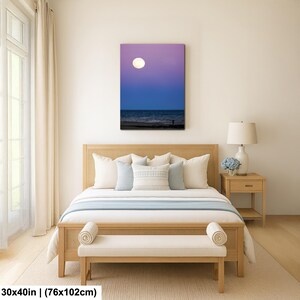 May include: A canvas print depicting a nighttime beach scene with a large, bright moon in a purple and blue sky. The artwork is displayed above a bed with light-colored bedding and a wooden frame. The print measures 30x40 inches.