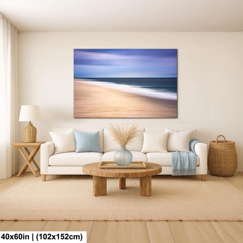 May include: A large canvas print depicting a beach scene with blurred motion. The artwork, measuring 40x60 inches, features a sandy shore, ocean waves, and a blue and purple sky. The print hangs above a white sofa in a living room setting.