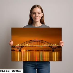 May include: A canvas print featuring a nighttime view of a bridge with illuminated lights reflecting in the water. The bridge has an arched design with a red light at the top. The print measures 24x36 inches.
