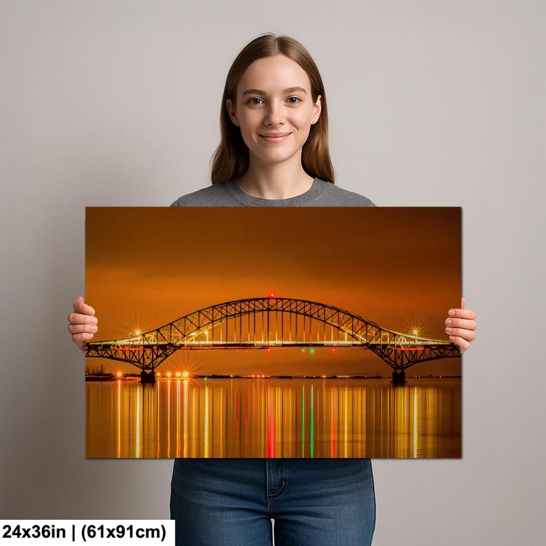 May include: A canvas print featuring a nighttime view of a bridge with illuminated lights reflecting in the water. The bridge has an arched design with a red light at the top. The print measures 24x36 inches.