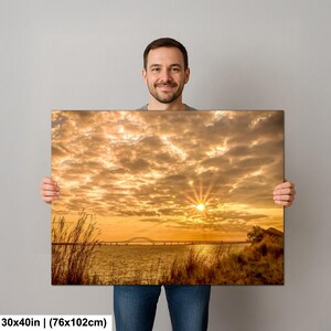May include: A canvas print featuring a vibrant sunset over a body of water, with a bridge in the distance. The sky is filled with golden clouds, and the sun is shining brightly. The print measures 30x40 inches.