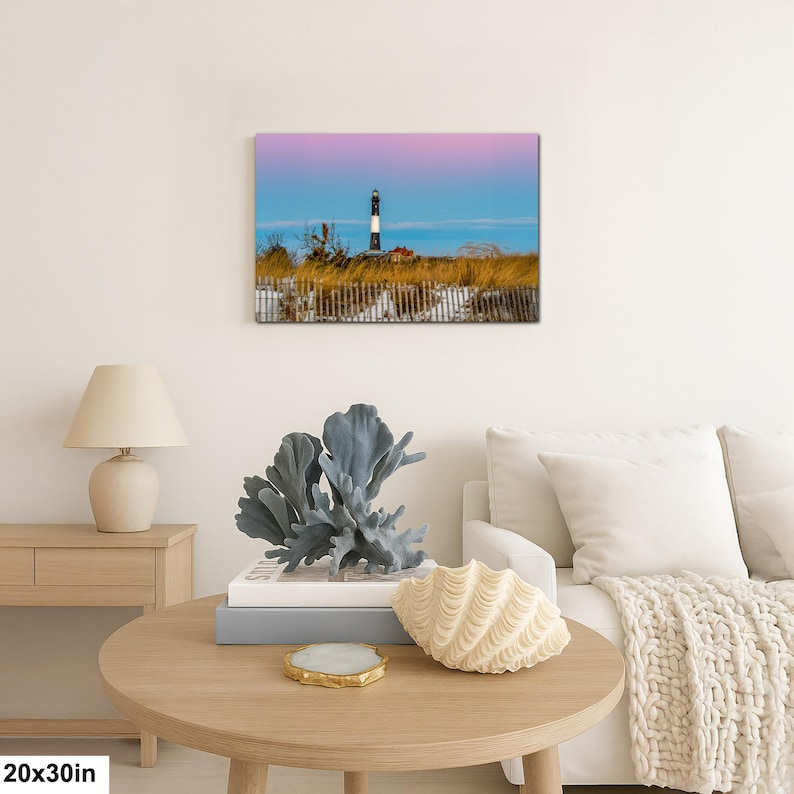 Op de afbeelding: Een 50x76 cm canvas print van een kustlandschap met een zwart-witte vuurtoren onder een kleurrijke lucht. De vuurtoren staat achter een houten hek en hoog gras. De print wordt weergegeven boven een ronde houten tafel met decoratieve items.