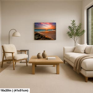 May include: A framed canvas print of a beach scene with a vibrant sunset. The artwork, measuring 16x20 inches, features a colorful sky, ocean waves, and a sandy shore. The print is displayed in a modern living room setting.