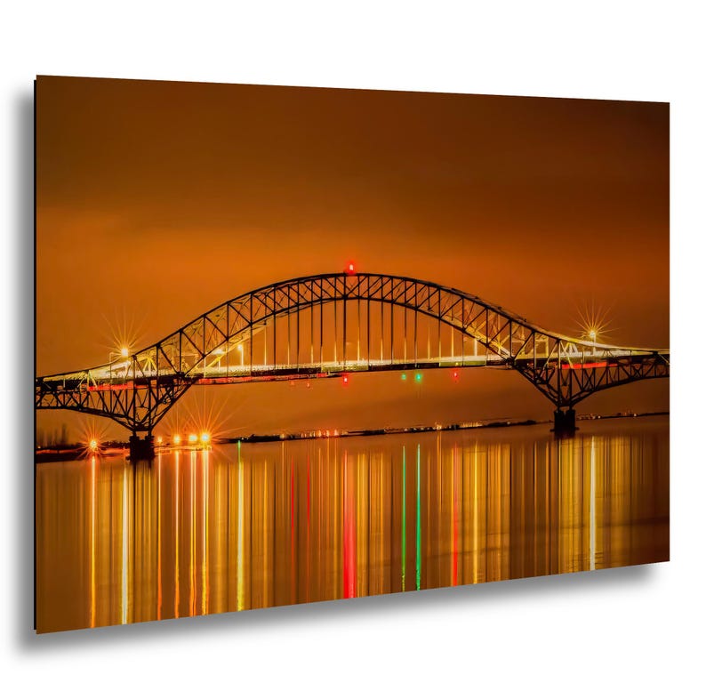 May include: A nighttime photograph of a bridge with a curved arch design. The bridge is illuminated with lights, reflecting in the water below. The sky is a warm orange color, and the overall scene is a vibrant display of light and color.