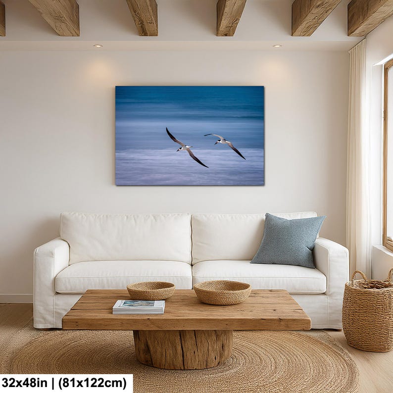 May include: A canvas print of two seabirds in flight over a blurred ocean. The artwork, measuring 32x48 inches, features a blue and white color palette, creating a serene coastal scene. The birds' wings are outstretched, adding a sense of movement.