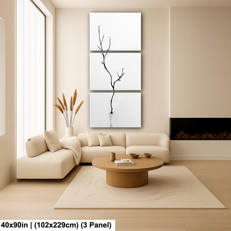 May include: A triptych canvas art piece featuring a minimalist black and white image of a slender tree branch. The artwork is displayed in a modern living room setting, with a neutral color palette and a large window.