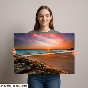 May include: A 24x36 inch (61x91cm) canvas print of a beach sunset. The image features a vibrant sunset with orange, pink, and blue hues over the ocean. A flock of birds flies across the sky, and the foreground shows a sandy beach and rocky shoreline.