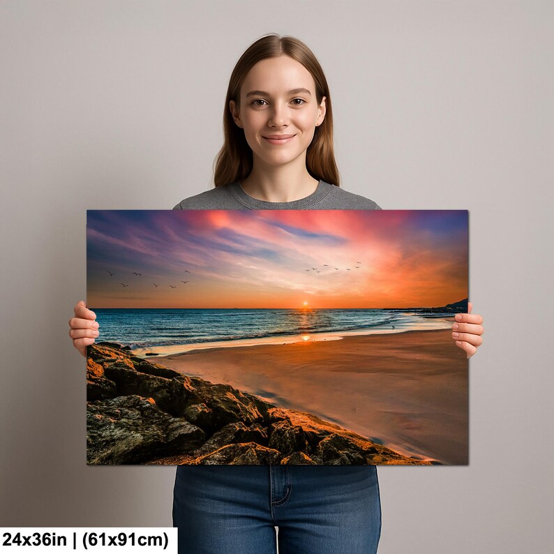 May include: A 24x36 inch (61x91cm) canvas print of a beach sunset. The image features a vibrant sunset with orange, pink, and blue hues over the ocean. A flock of birds flies across the sky, and the foreground shows a sandy beach and rocky shoreline.